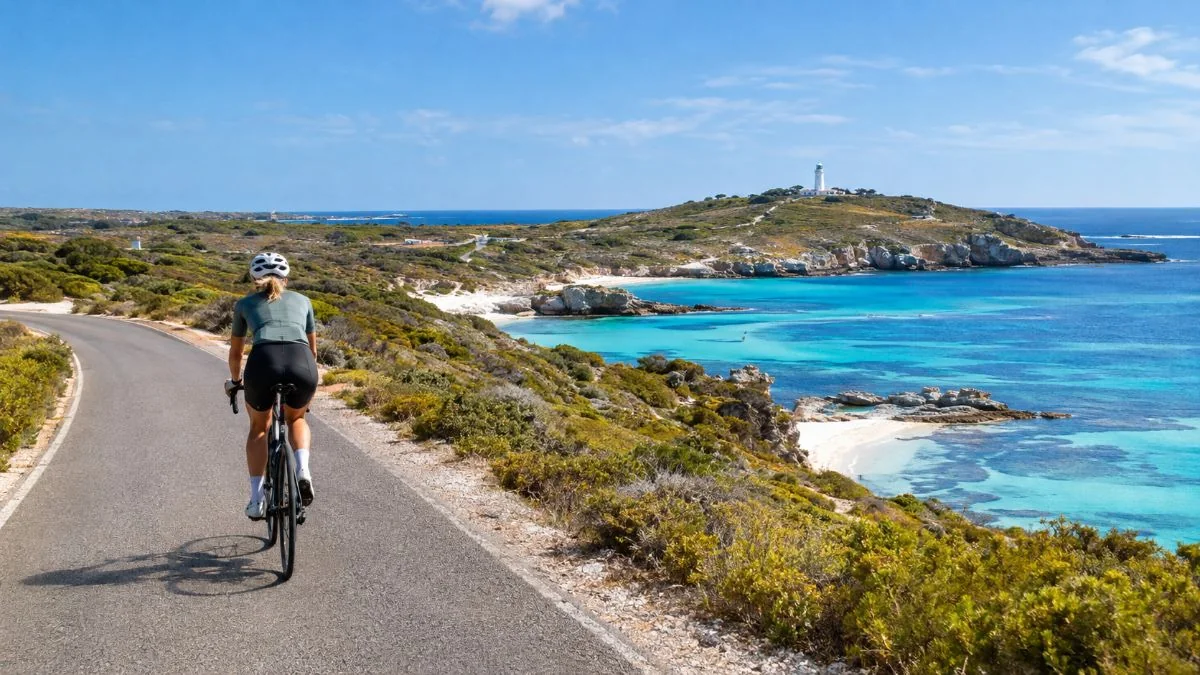 A cyclist riding a quiet coastal road above turquoise bays with a lighthouse on a distant hill