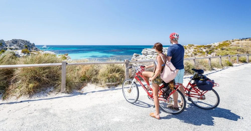 Two cyclists with red hire bikes at a lookout over a turquoise bay on Rottnest Island